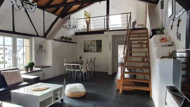 Ampla sala em estilo loft com mesa de jantar, cadeiras, sofá, mesa de centro branca e escada para um andar superior.
