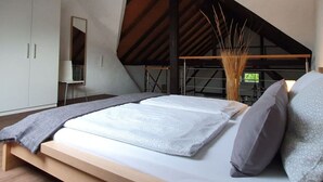 Quarto com teto inclinado, cama com manta cinza, travesseiro branco e cabeceira de madeira.