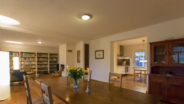 A spacious living room with a wooden dining table, chairs, a bookshelf, and a kitchen area. - in the Estate Worker's House (Karstädt)
