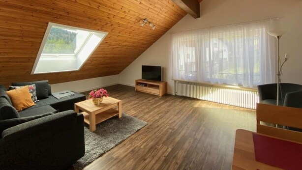 A loft-style room with a sloped ceiling, wooden beams, a sofa, a coffee table, a TV, and a window with a view of greenery. - Spacious Holiday Home Near Ski-lift (Todtmoos)