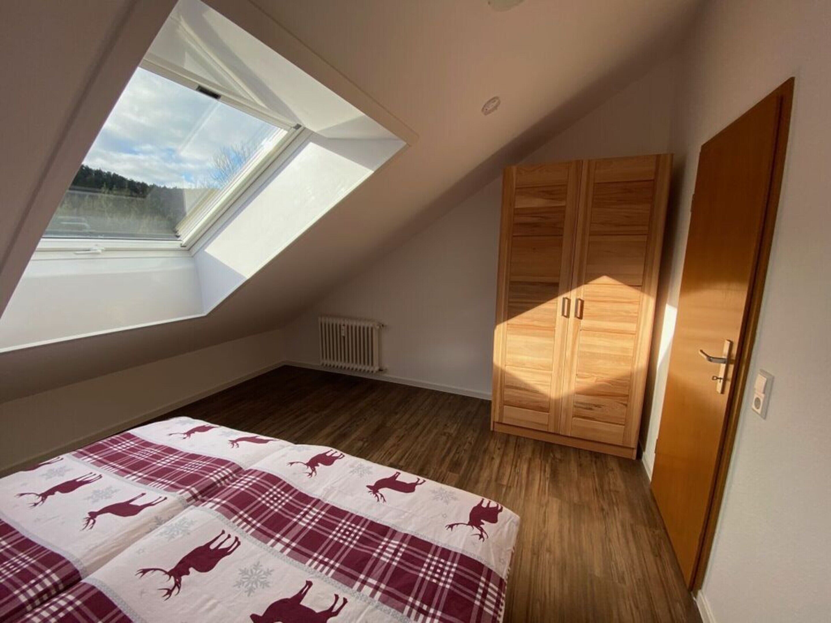 A bedroom with a sloped ceiling, a skylight, a bed with a plaid bedspread, wooden wardrobes, and a radiator.