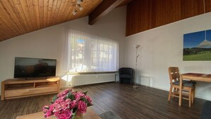 A spacious living room with a wooden ceiling, a large window with sheer curtains, a flat-screen TV, a wooden coffee table with a vase of flowers, a floor lamp, and a painting on the wall. - Spacious Holiday Home Near Ski-lift (Todtmoos)
