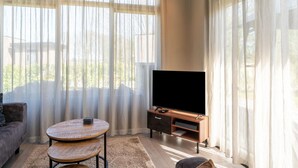 Ein Wohnzimmer mit einer Couch, einem runden Holztisch, einem Fernseher auf einem Holzschrank und transparenten Vorhängen.