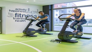 Two people using stationary bikes in a gym with a wall displaying words related to fitness and health. - Comfortable Apartment With air Conditioning (Bruges)