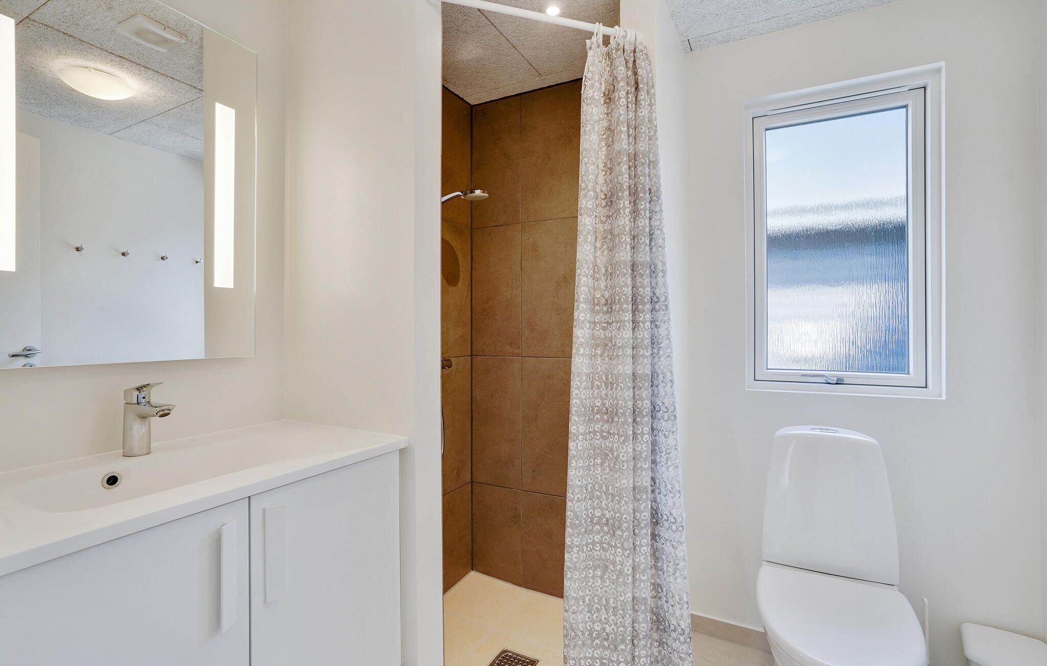 A bathroom with a white sink, mirror, and toilet, a shower with a patterned curtain, and a window with a view of a snowy landscape.