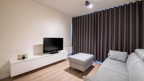 Un salon moderne avec un téléviseur à écran plat posé sur une console blanche, un canapé gris et une grande fenêtre ornée de rideaux foncés.