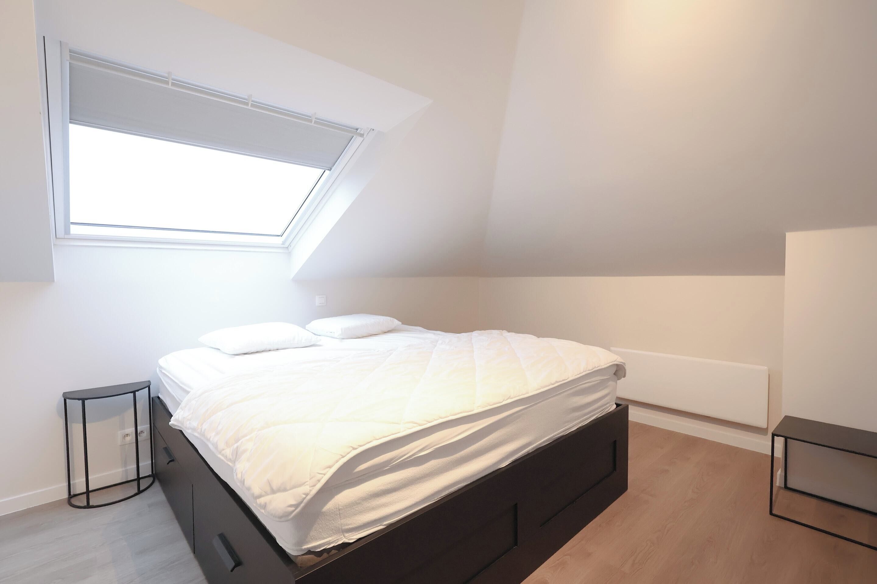 A bedroom with a skylight, a bed with white bedding, a small bedside table, and a radiator.