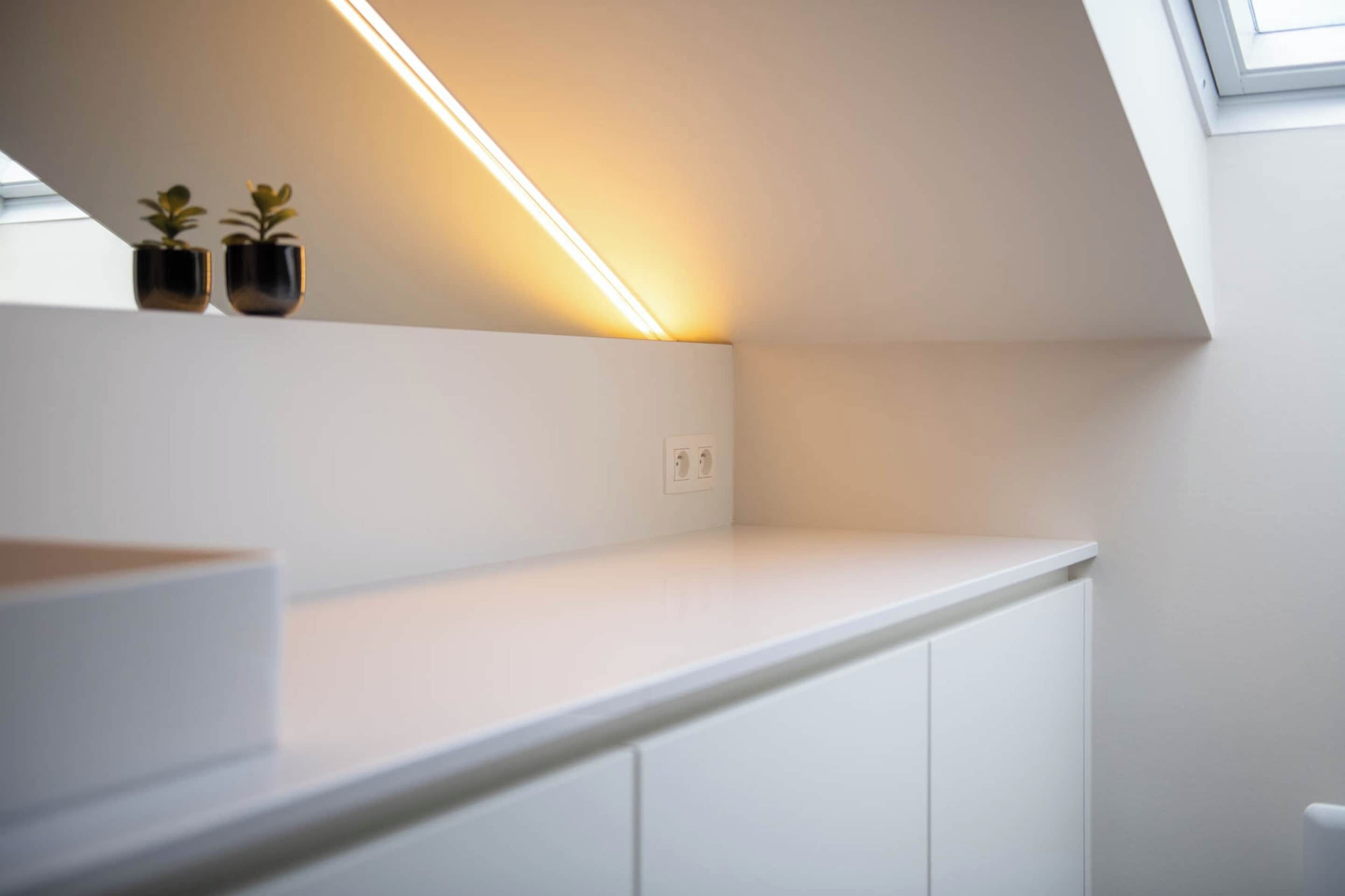 A modern, white interior with a slanted ceiling, a built-in shelf with two potted plants, and a visible electrical outlet.