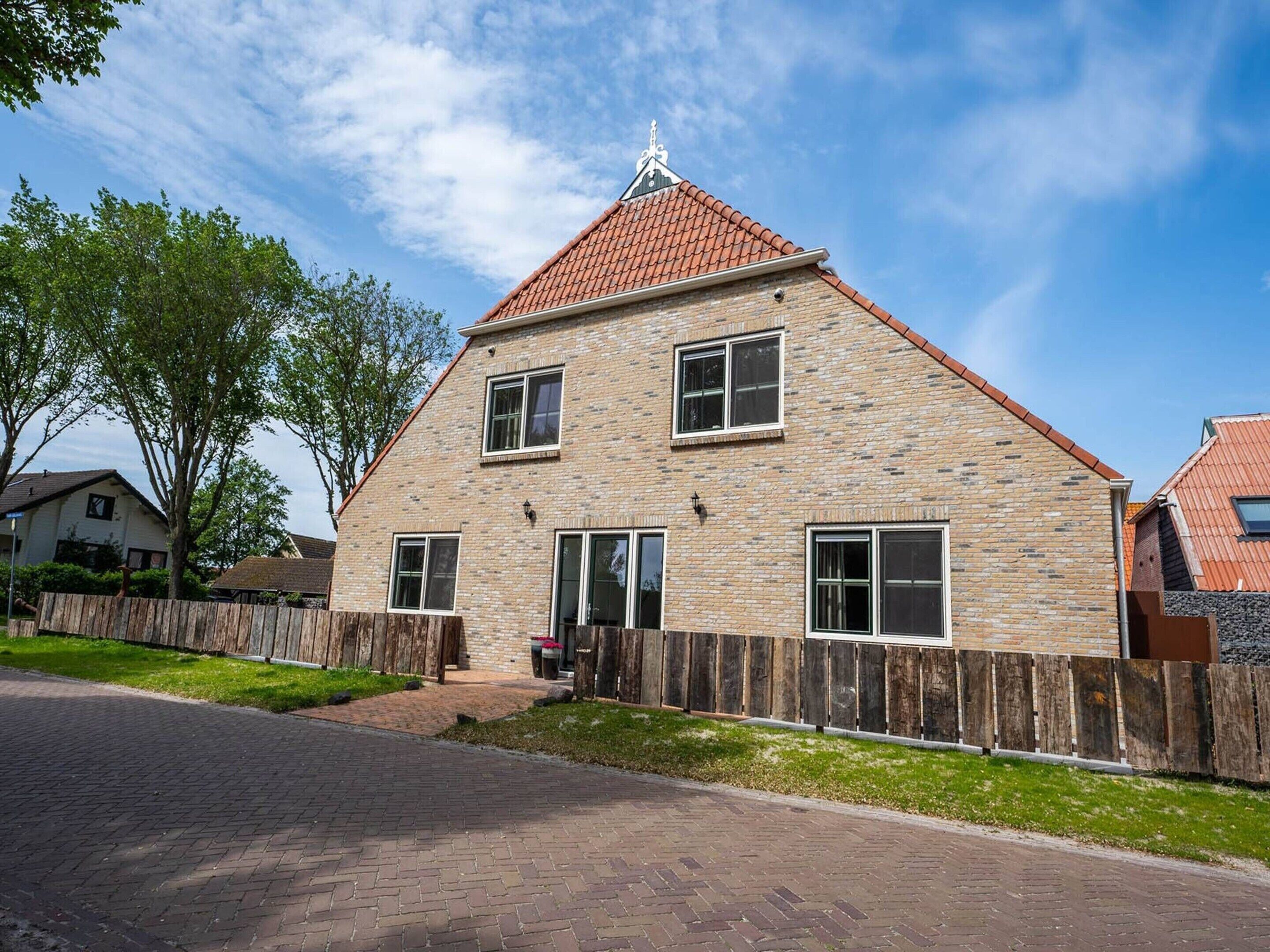 Une maison à deux étages, construite en briques, avec un toit recouvert de tuiles rouges, une clôture en bois et une allée pavée.