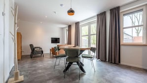 Ein geräumiger Essbereich mit Holztisch und -stühlen, einem an der Wand befestigten Fernseher und großen Fenstern mit Vorhängen.