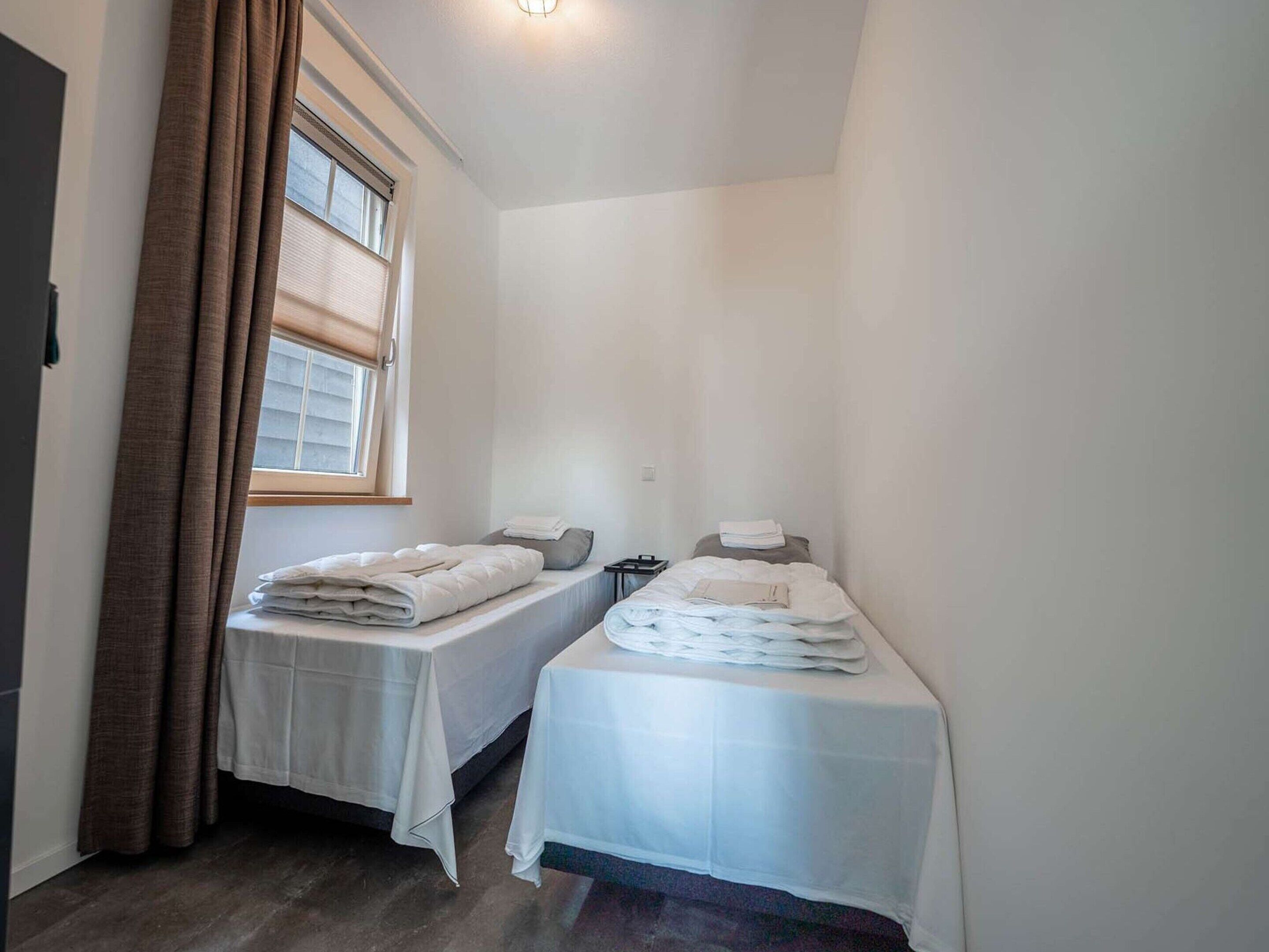 A small, clean room with two beds, a window with blinds, and a bedside table with a lamp.