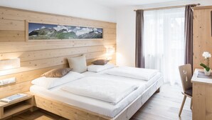 A modern bedroom with a wooden bed, a nightstand, a desk, and a large window with curtains. - Apartment in St. Jakob im Ahrntal Sudtirol (Valle Aurina)