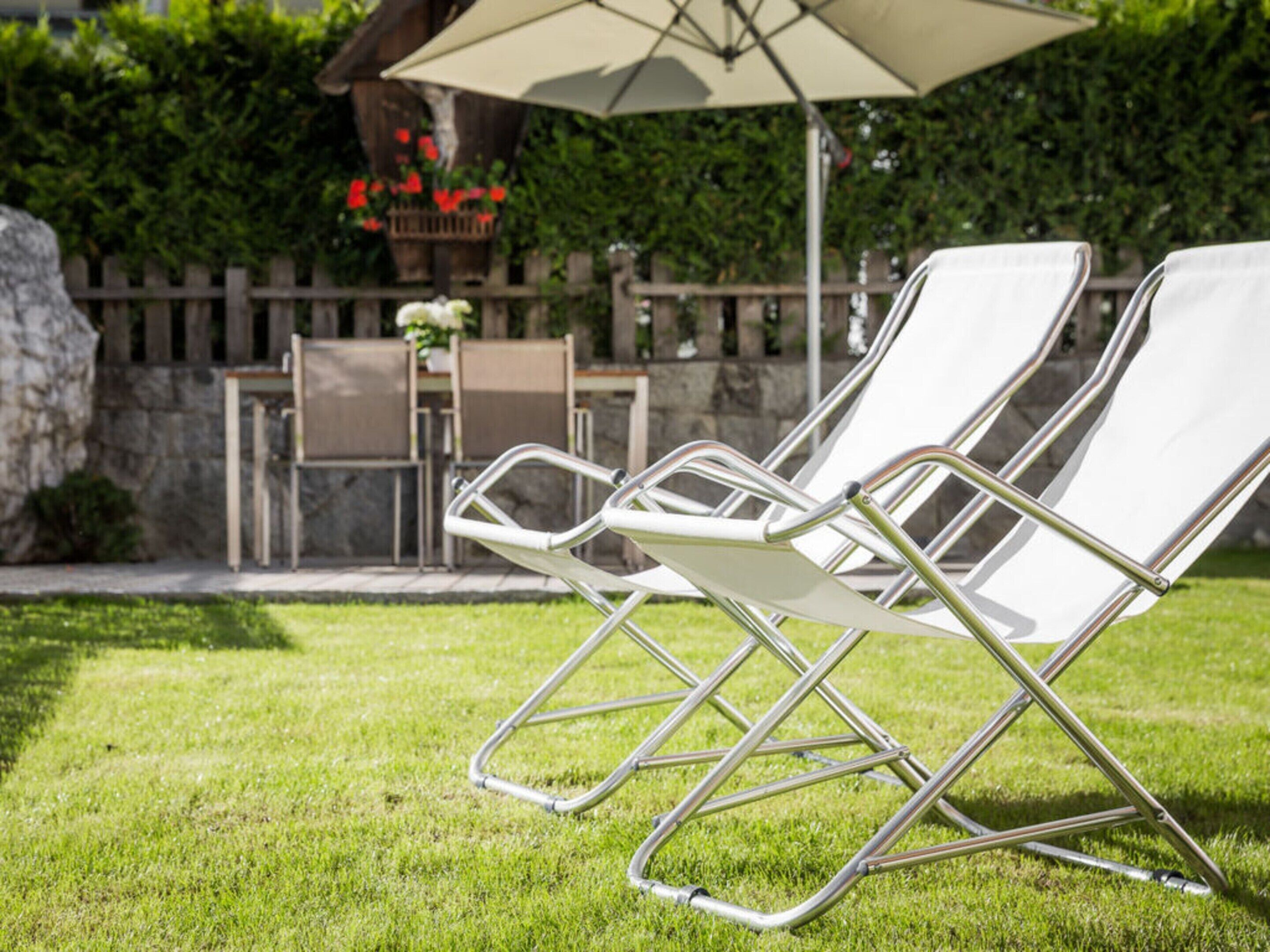 Zwei weiße Lounge-Stühle mit Metallgestellen, ein Tisch und ein Sonnenschirm in einem Garten.