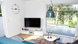 A modern living room with a flat-screen TV, a blue sofa, a small coffee table, and a large window with a view of a wooden deck. - Holiday Apartment Elbe (Drochtersen)