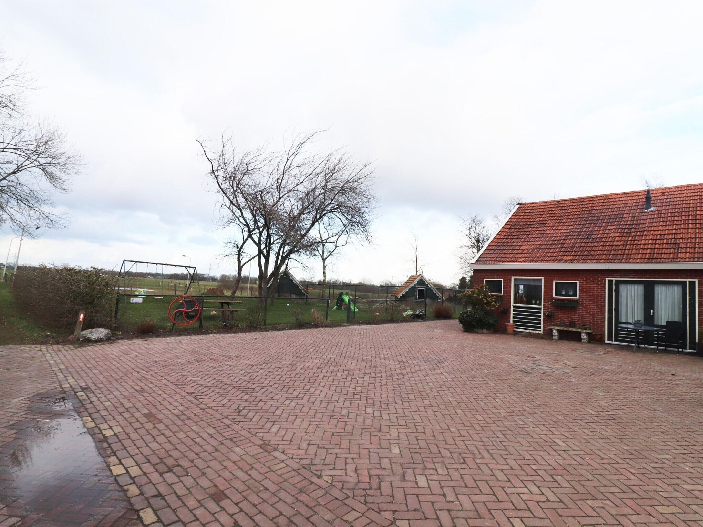 A brick-paved courtyard with a red-roofed building, a swing set, and a fenced area with trees.