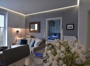 A modern living room with a sofa, a dining table with plates and cutlery, a bed with blue bedding, and a wall-mounted mirror. - Watt`n Flair on the North Beach (Borkum)