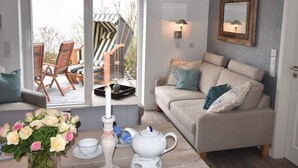 A cozy living room with a sofa, a table with a vase of flowers, and a view of outdoor furniture through a window. - Watt`n Flair on the North Beach (Borkum)
