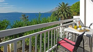 Een balkon met een witte railing, een tafel met een rode stoel, en uitzicht op zee en bergen.