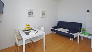 Een moderne woonkamer met een witte tafel, een blauwe bank en een kleine witte tafel met een groene map. Twee ingelijste foto's hangen aan de muur en er is een plafondlamp.
