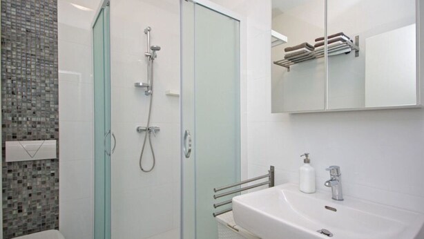Een badkamer met een douchecabine van glas, een wit wastafelmeubel met zeepdispenser en een betegelde wand met een mozaïekpatroon.