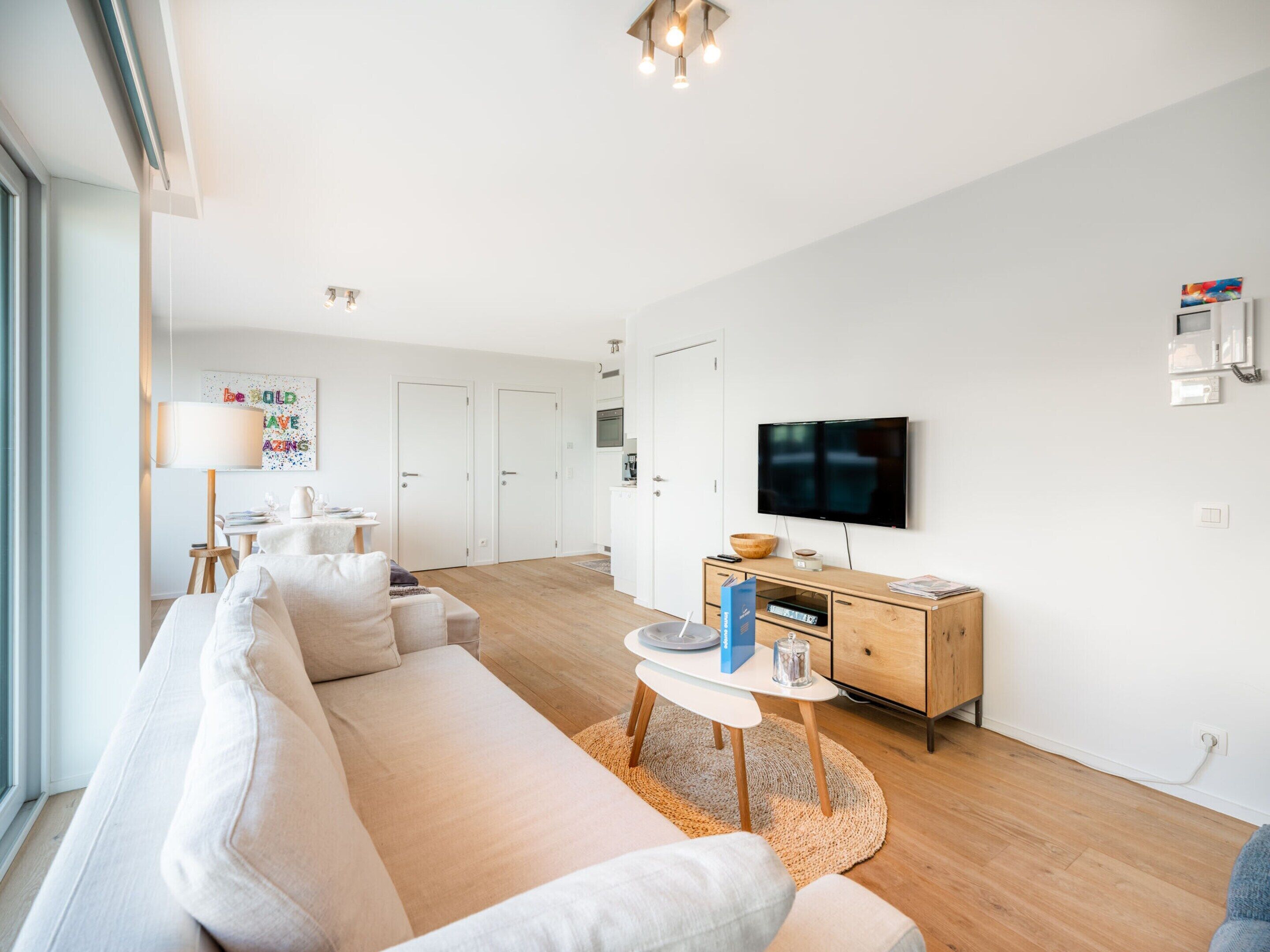 A modern living room with a sofa, coffee table, and TV. There are white walls, wooden flooring, and a large window.