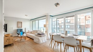 A modern living room with a sofa, dining table, and chairs, large windows with a view of buildings, and a flat-screen TV mounted on the wall. - Apartment in Koksijde With Sea Views (Koksijde)