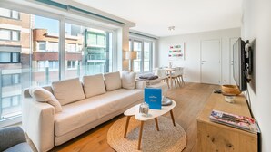 A modern living room with a large window, a sofa, a coffee table, and a dining area. - Apartment in Koksijde With Sea Views (Koksijde)