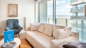 A modern living room with a beige sofa, a blue armchair, and a large window overlooking a beach. - Apartment in Koksijde With Sea Views (Koksijde)