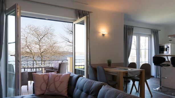 A modern living room with a sofa, dining table, chairs, and a view of a tree through large windows. - Casa sul Lago Modern Retreat (Werder (Havel))