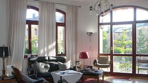 Een ruime woonkamer met een groot raam, een zwarte leren bank, een rode fauteuil en een kroonluchter.