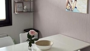 A dining area with a white table, white chairs, a white hanging light fixture, a framed picture of a beach scene, and a small shelf with decorative items. - Holiday Home Meerhuus (Butjadingen)
