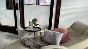 A sun lounger with a fur throw and a pillow with text, a small table with a vase and a sign, and a view of a building through a window. - Holiday Home Meerhuus (Butjadingen)