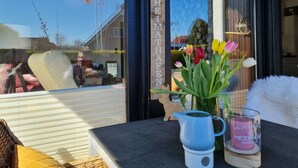A view through a window with a sign reading "HEIMHAFEN," a blue kettle, a glass candle holder, and a vase with tulips. - Holiday Home Meerhuus (Butjadingen)