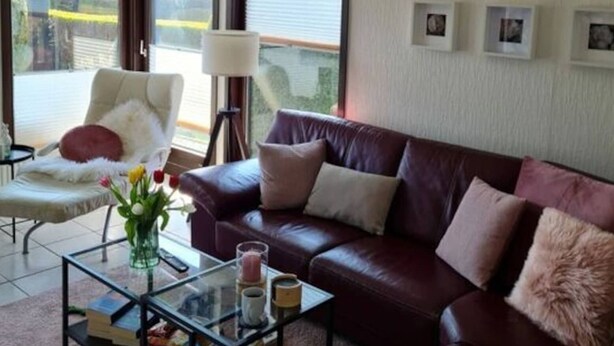 A living room with a leather sofa, a glass coffee table, a white armchair, and a large window with blinds. - Holiday Home Meerhuus (Butjadingen)