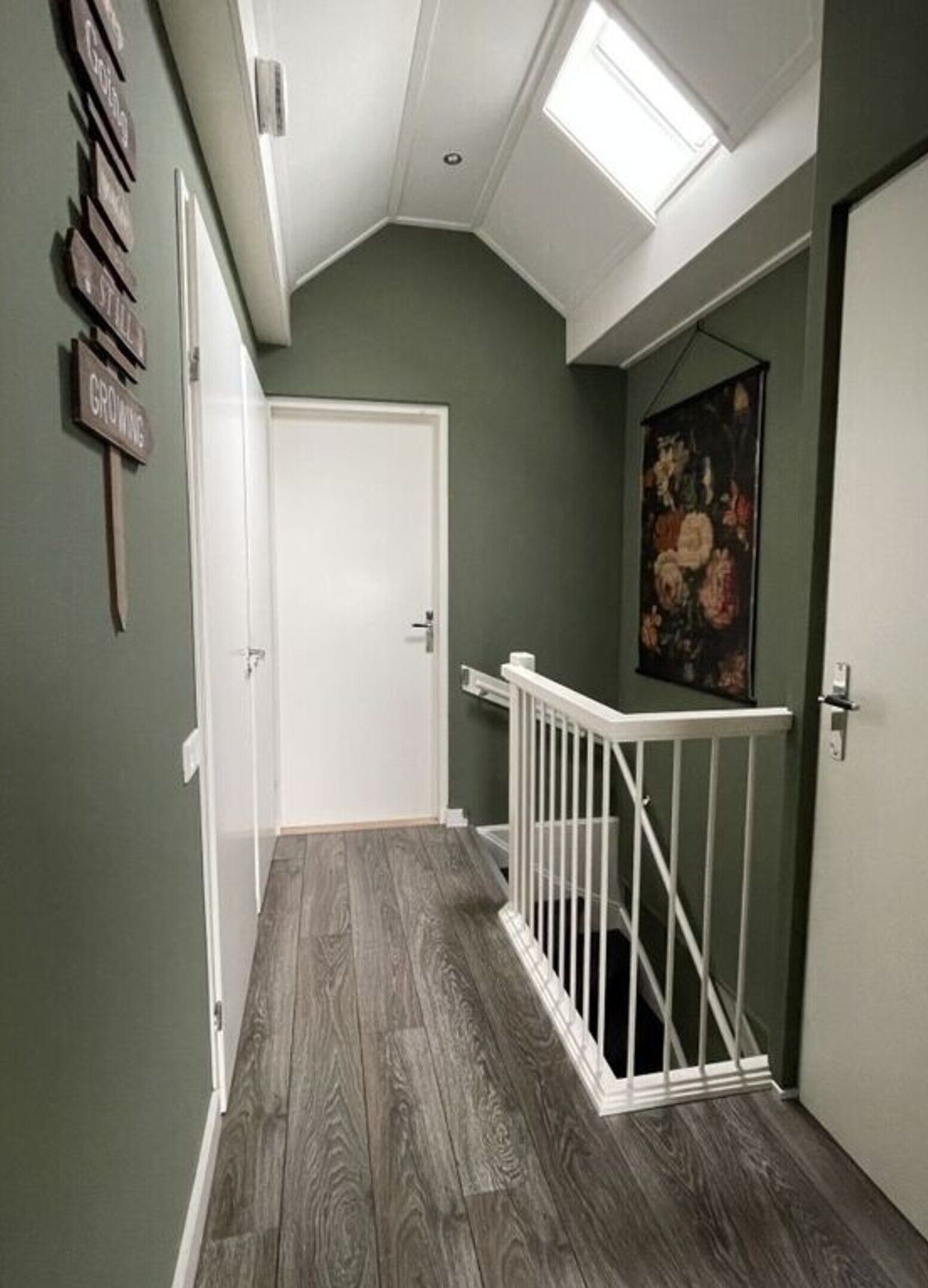 Un couloir avec un plafond en pente, une porte blanche, une rambarde blanche et un tableau accroché au mur.