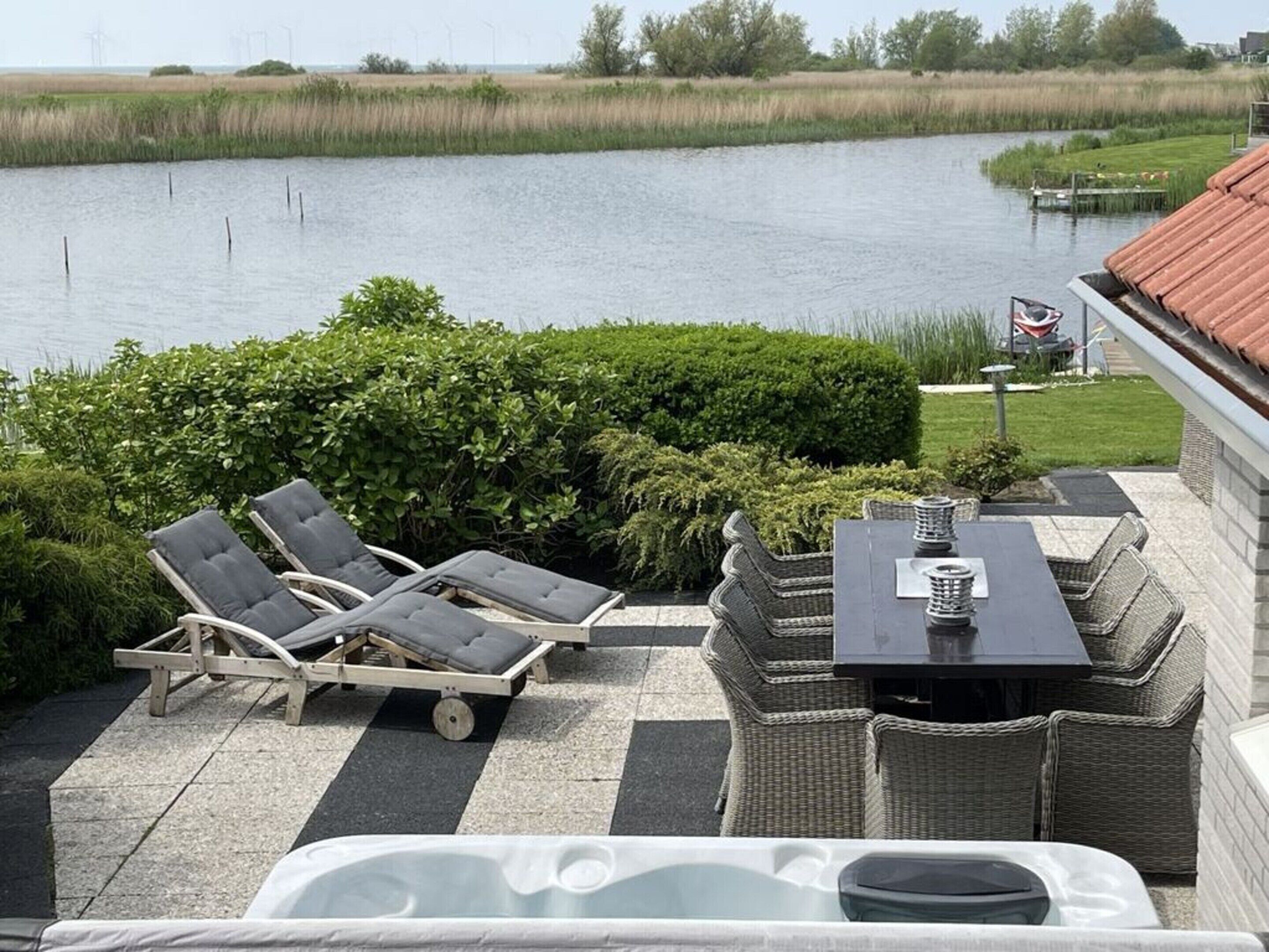 Een terras met lounge fauteuils en een tafel, met uitzicht op een wateroppervlak, en windturbines in de verte.