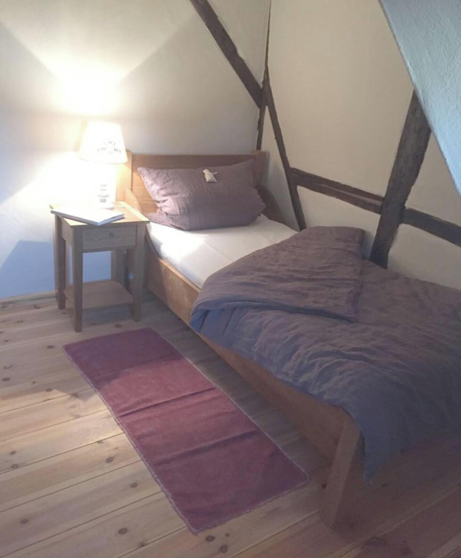 A bedroom with a wooden floor, a bed with a blue blanket, a wooden nightstand with a lamp, and a red rug.