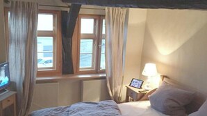 Ein Schlafzimmer mit einem Bett, einem Schreibtisch mit Computer, einem Fenster mit Vorhängen und einem Nachttisch mit Lampe.