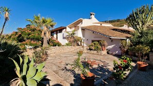 A white villa with a tiled roof, surrounded by a garden with various plants and a stone pathway.