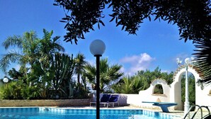 A swimming pool with a clear blue sky, surrounded by palm trees and a white building with an arched entrance.