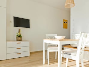 A modern, minimalist dining area with a wooden table and white chairs, a small potted plant, and a flat-screen TV mounted on the wall. - House in Reed 1 - Apartment 2 (Neukieritzsch)