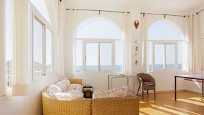 A room with a curved wicker sofa, a small table, a chair, and large arched windows overlooking the sea. - Belvilla by OYO Chalet Corralete (Nijar)