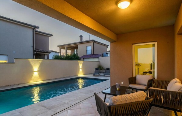 Un espace piscine avec une table et des chaises, un mur bien éclairé et une vue sur les bâtiments voisins.