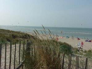 A beach with a wooden fence and tall grass in the foreground, a sandy beach with people and tents in the middle ground, and the sea with kites in the background. - Strandjutter Modern Retreat (Vrouwenpolder)