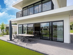 Une maison moderne à deux étages, dotée d’un balcon, d’un coin repas avec une table et des chaises, et de grandes portes vitrées donnant sur l’intérieur.