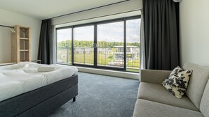 A hotel room with a bed, a sofa, a wooden wardrobe, and a large window with a view of greenery and buildings.