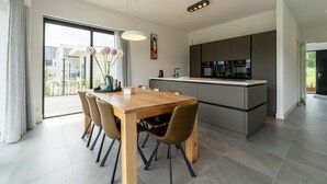 A modern kitchen with a central island, a dining area with a wooden table and chairs, and large windows offering a view of the outdoors.