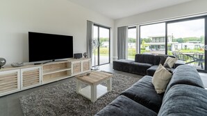 A modern living room with a flat-screen TV, a wooden coffee table, a grey sofa, and a large window offering a view of a modern building and greenery.