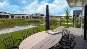 A modern outdoor patio with a wooden table and chairs, a view of a lake, and a row of contemporary buildings in the background.