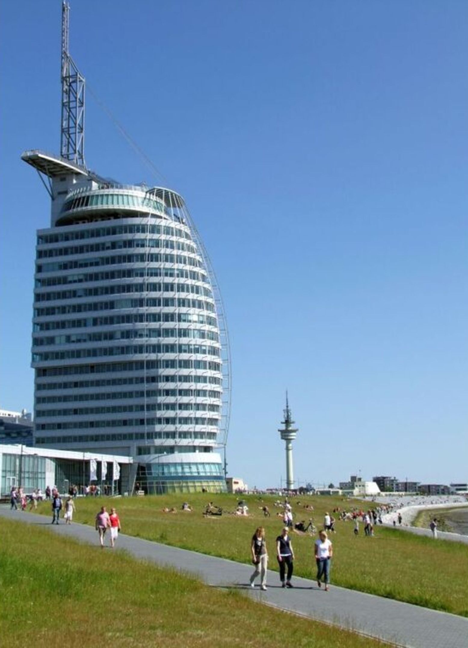 Ein modernes Gebäude mit gewölbter Fassade, einem markanten Turm und einer umliegenden Parkanlage mit Spaziergängern.